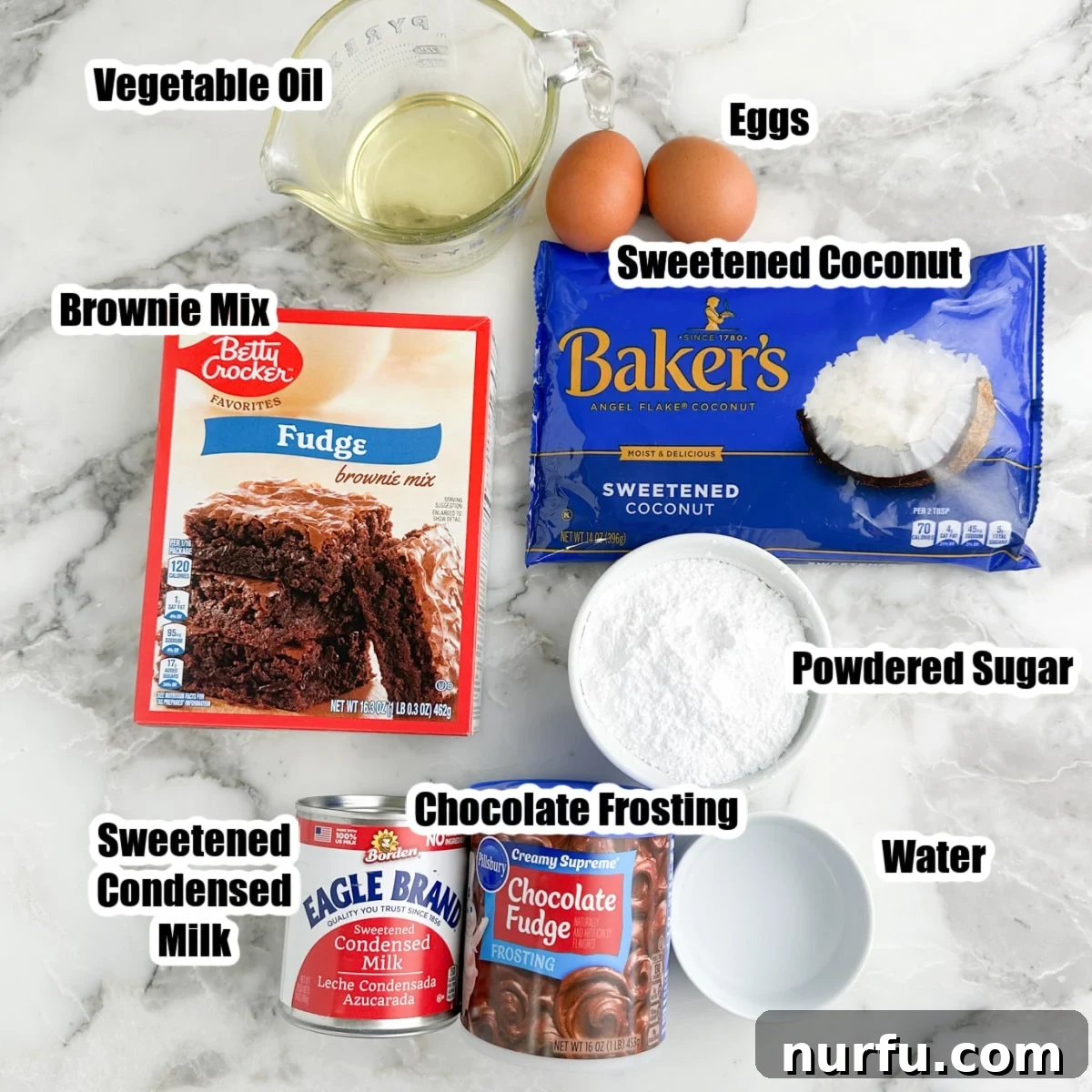 A flat lay photograph displaying all the key ingredients for coconut brownies: a box of brownie mix, eggs, vegetable oil, water, a bag of sweetened shredded coconut, sweetened condensed milk, powdered sugar, and a tub of chocolate frosting.