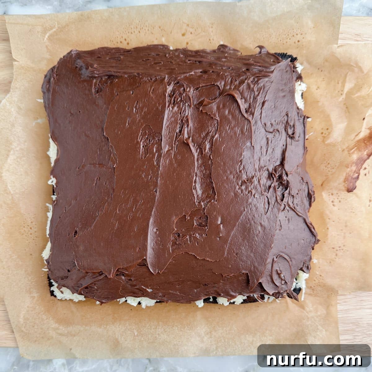 The final layer of smooth, melted chocolate frosting being spread over the coconut-topped brownies, creating a glossy, enticing finish.