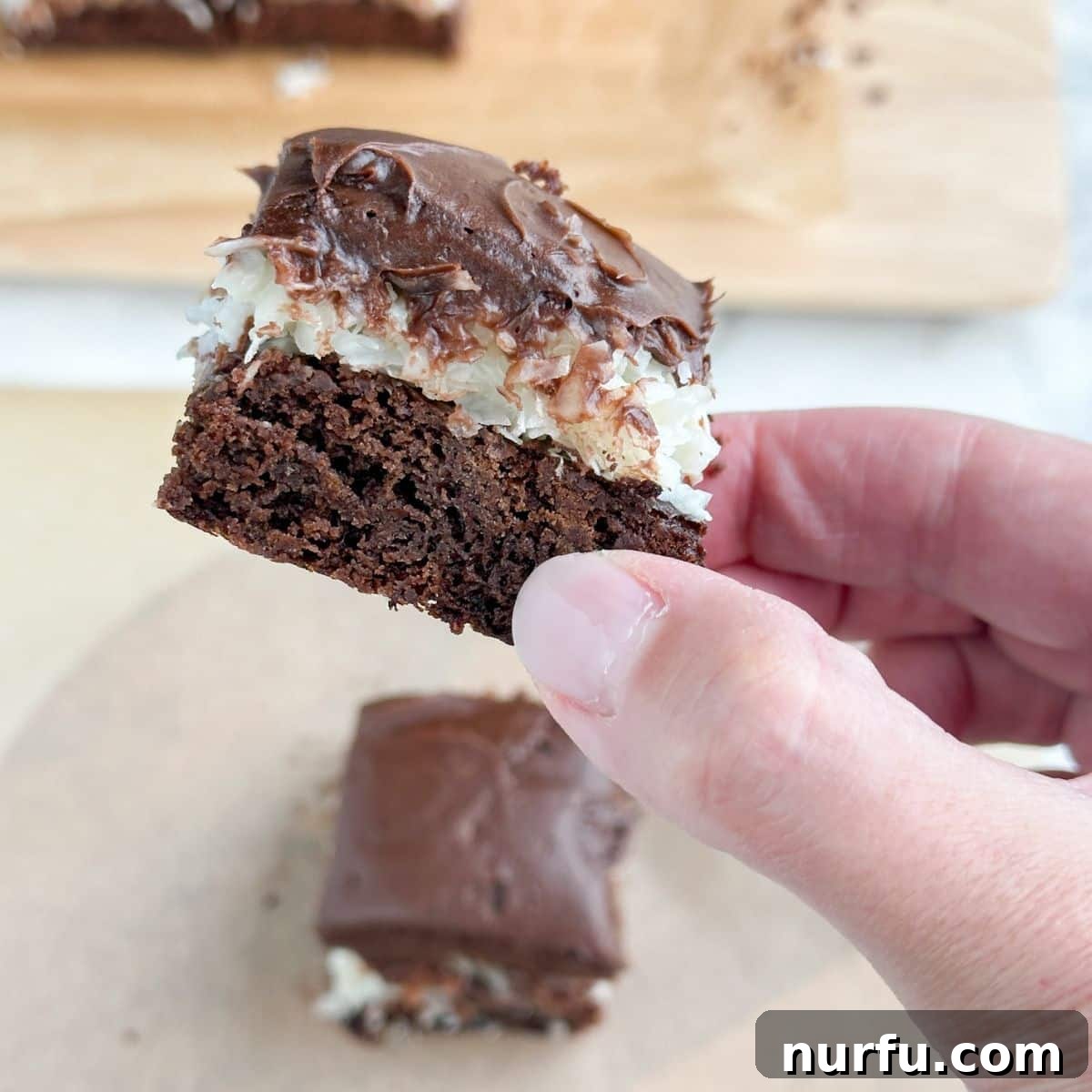 A hand reaching for a perfectly cut coconut brownie, highlighting the thick, enticing layers and appealing texture.