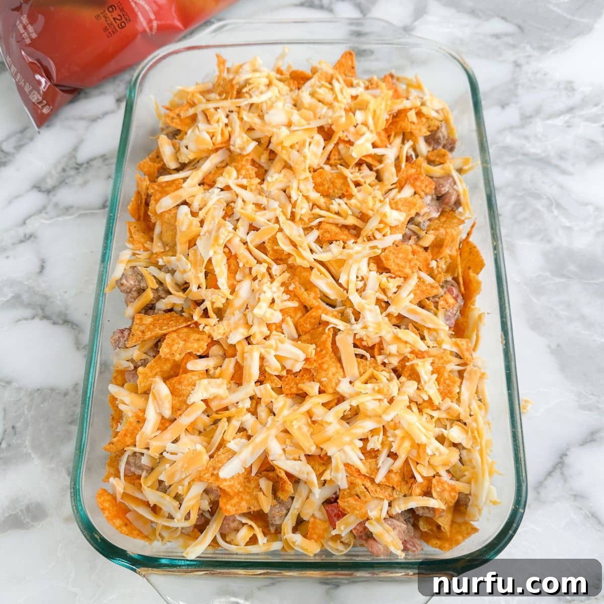 Casserole dish with all layers of unbaked Doritos casserole, topped with chips and cheese.