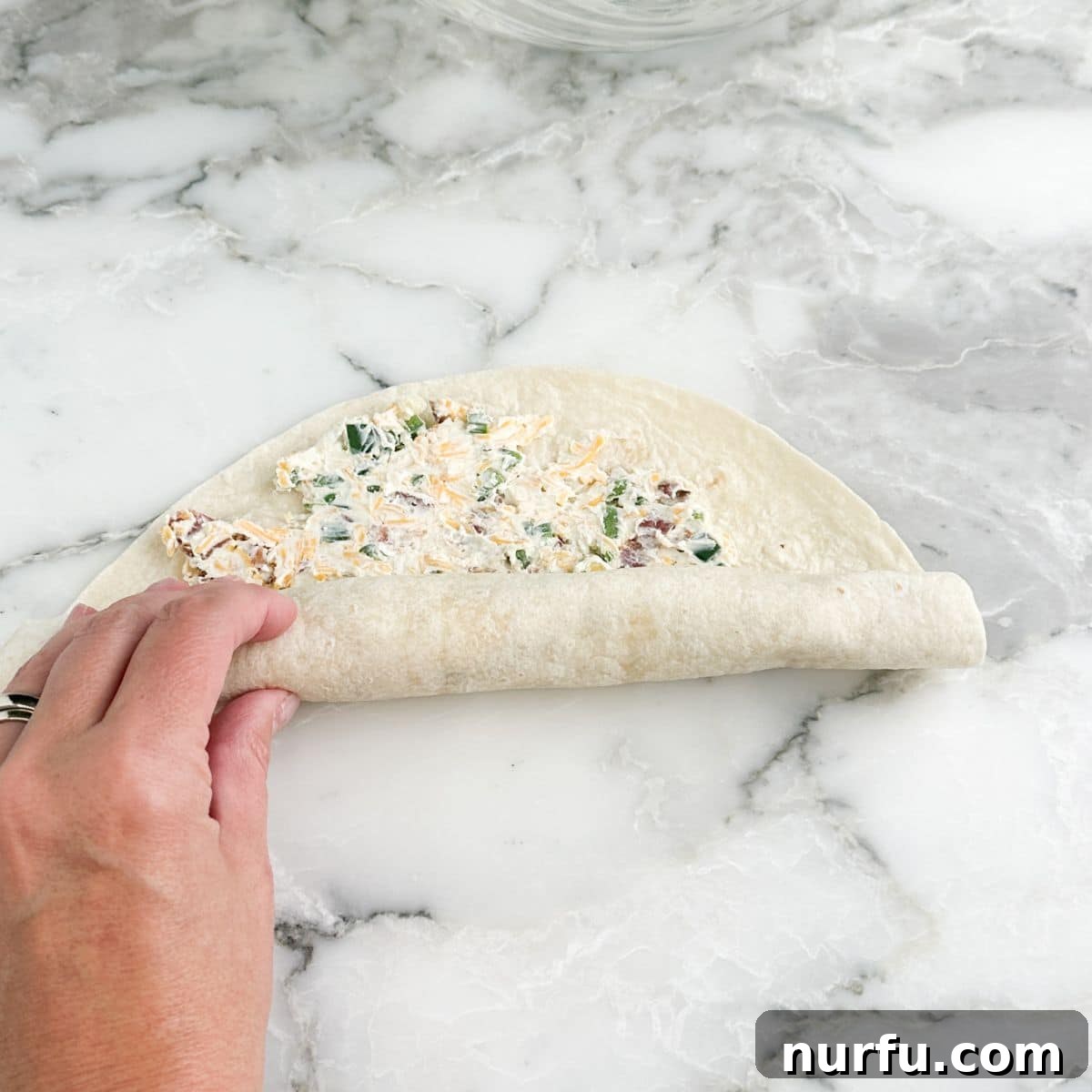 Cheesy Jalapeno Swirls 7 A hand carefully rolling up a flour tortilla filled with the jalapeño popper mixture, demonstrating the essential tight rolling technique for perfect pinwheels.