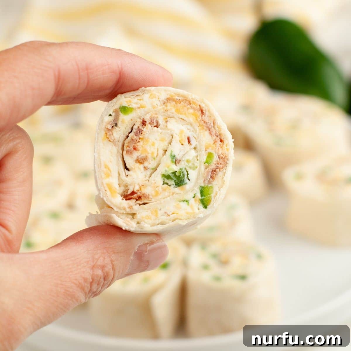 Cheesy Jalapeno Swirls 8 A hand gently holding a single, perfectly sliced Jalapeño Popper Pinwheel, highlighting its appealing spiral and delicious filling, against a blurred background.