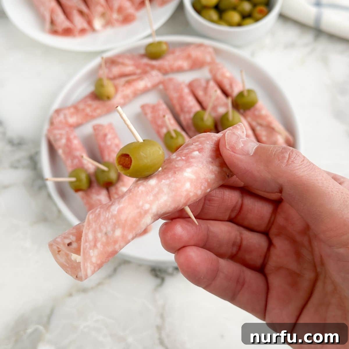 Savory Salami Bites 8 A hand holding a perfectly rolled salami roll up with a green olive on a toothpick.