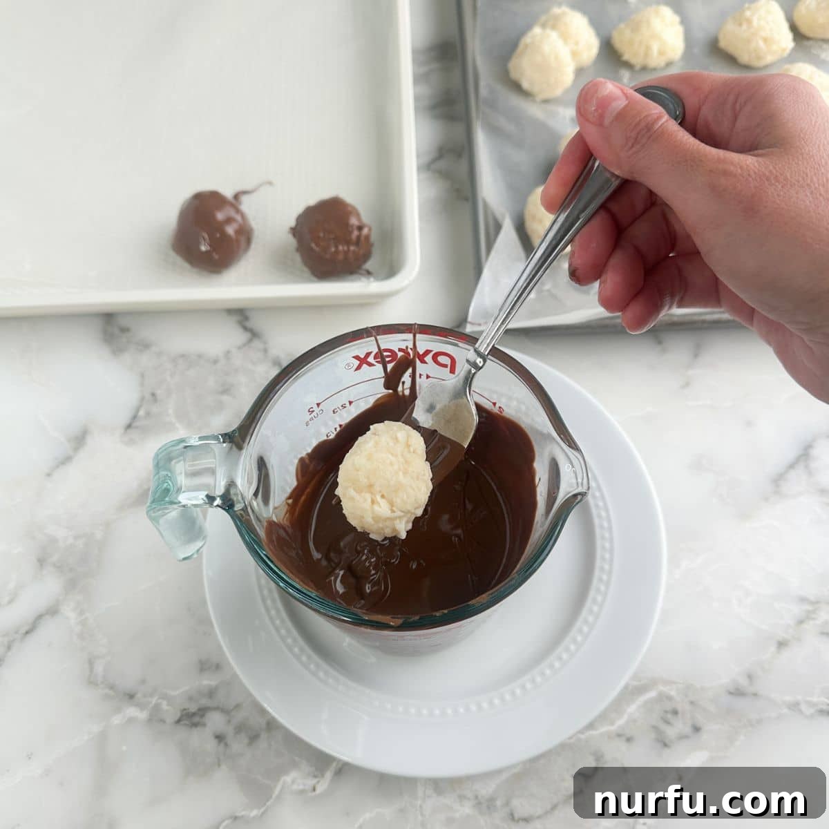 Coconut Chocolate Dream Bites 7 A coconut ball being carefully dipped into a bowl of melted chocolate with a fork, showing the smooth, even coating.
