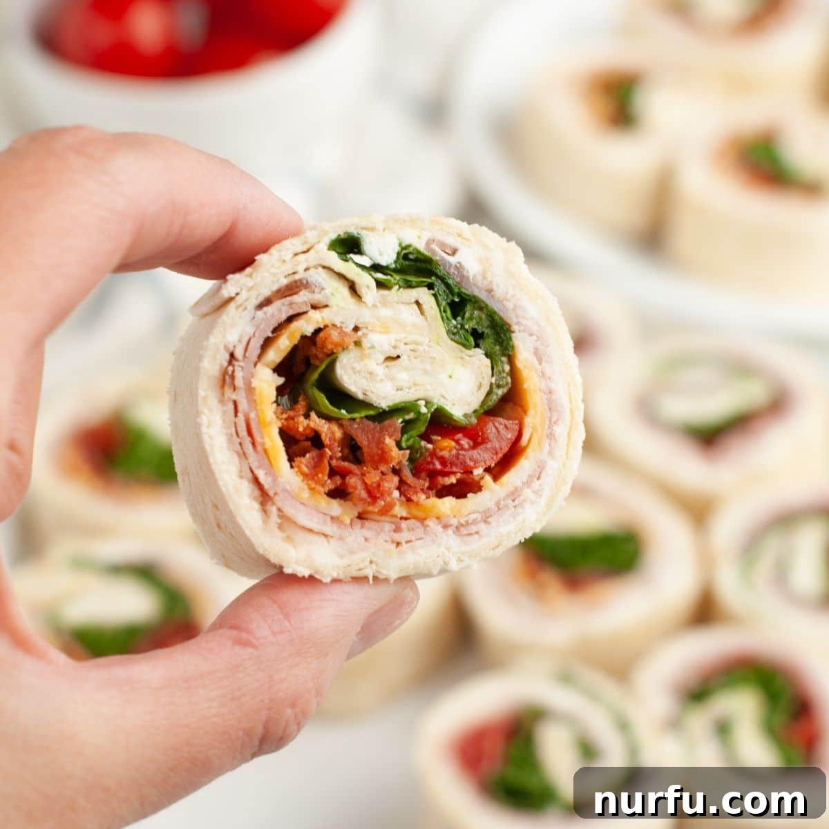 Clubhouse Pinwheel Bites 2 A hand holding a perfectly sliced club sandwich pinwheel, showcasing layers of turkey, ham, bacon, cheese, lettuce, and tomato wrapped in a tortilla.