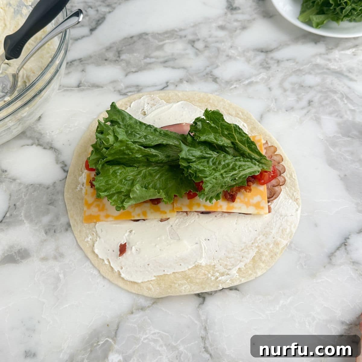 Clubhouse Pinwheel Bites 7 The tortilla, now complete with creamy spread, deli meats, cheese, bacon, and tomatoes, with fresh green lettuce leaves placed along the center.