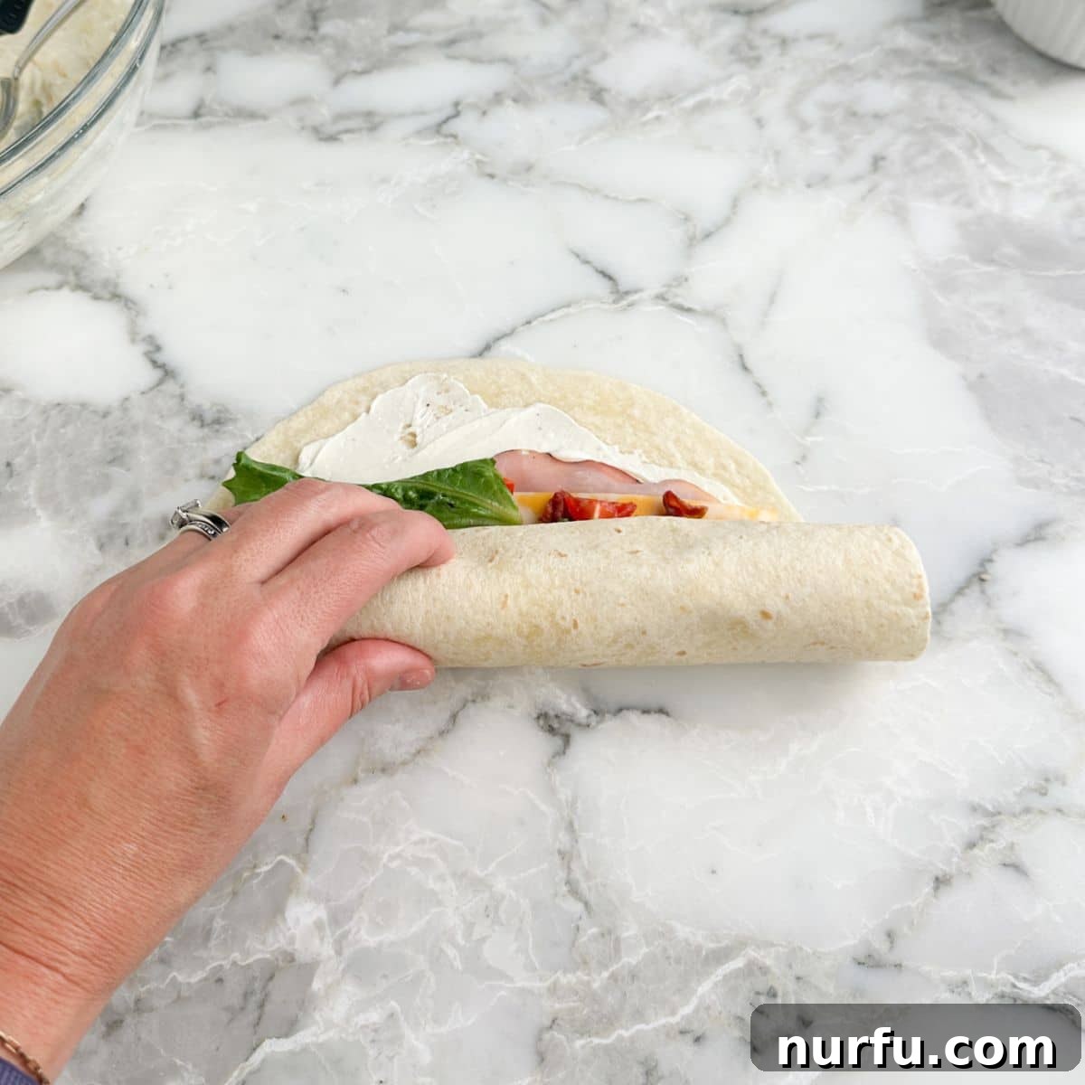 Clubhouse Pinwheel Bites 8 A hand carefully rolling up a fully loaded club sandwich tortilla lengthwise, ensuring all ingredients are tightly enclosed.