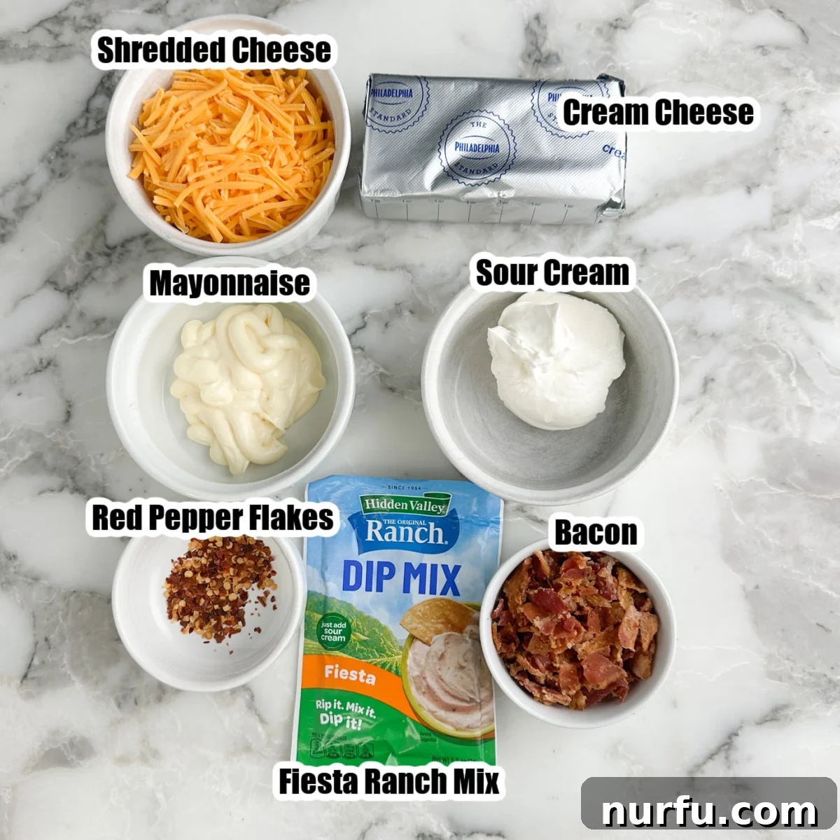 A collection of ingredients laid out for Fire Cracker Dip: a packet of Fiesta Ranch seasoning, bowls of shredded cheese, creamy mayonnaise, tangy sour cream, softened cream cheese, crispy crumbled bacon, and fiery red pepper flakes.
