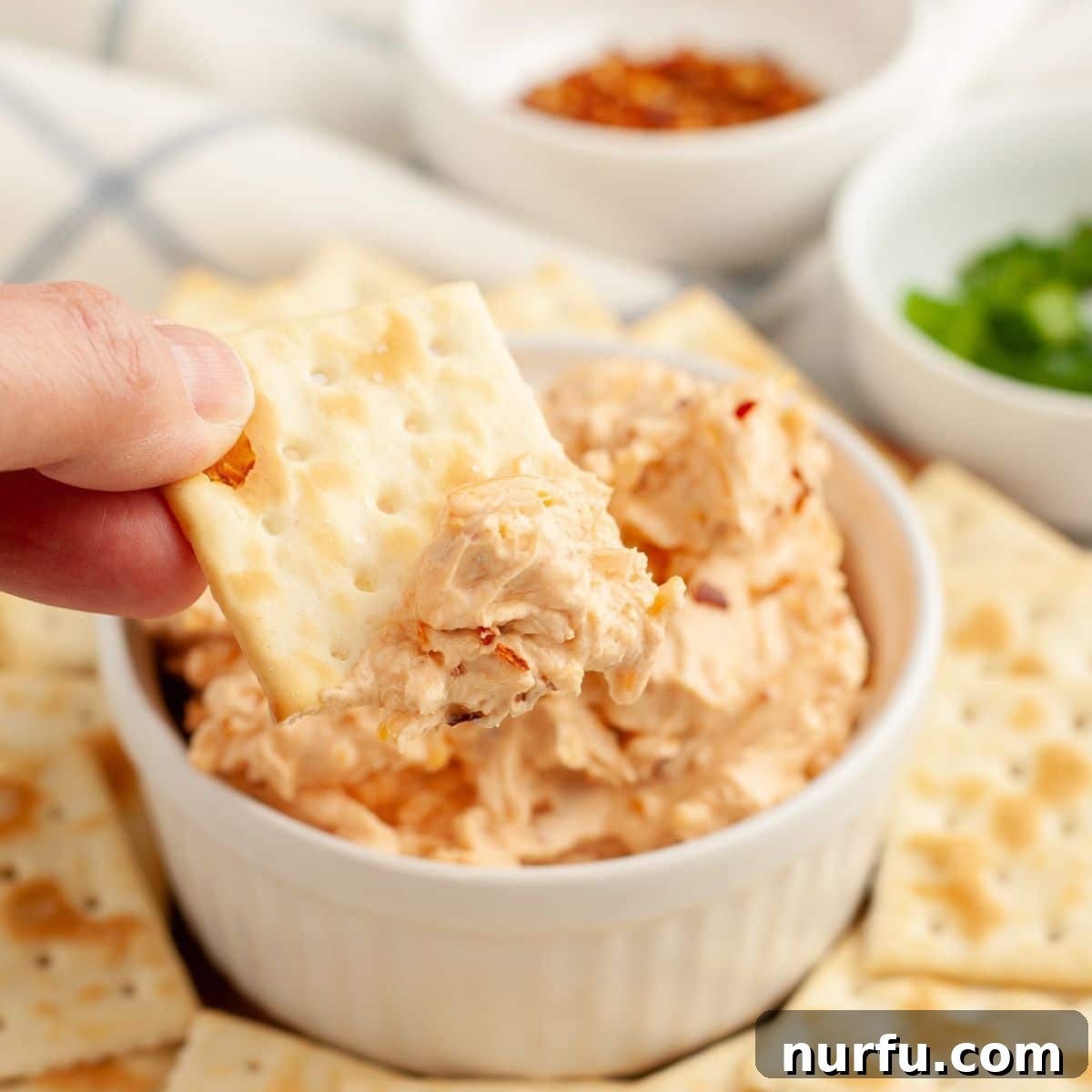A hand reaching for a cracker to dip into a bowl of Fire Cracker Dip, showcasing its inviting texture and readiness for enjoyment.