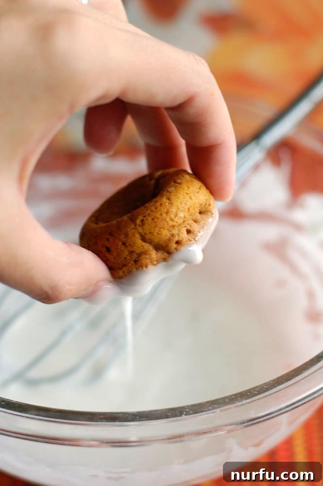 A mini pumpkin donut being dipped into a bowl of vanilla glaze.