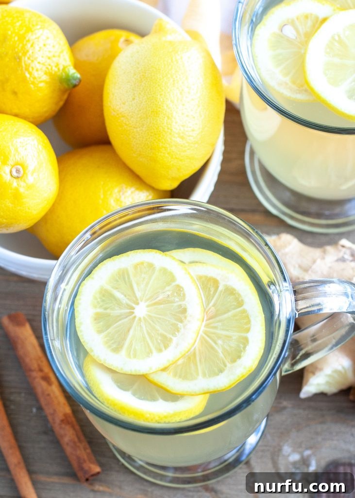 A glass of amber-colored ginger tea, garnished with lemon wedges, alongside a bowl of fresh lemons.