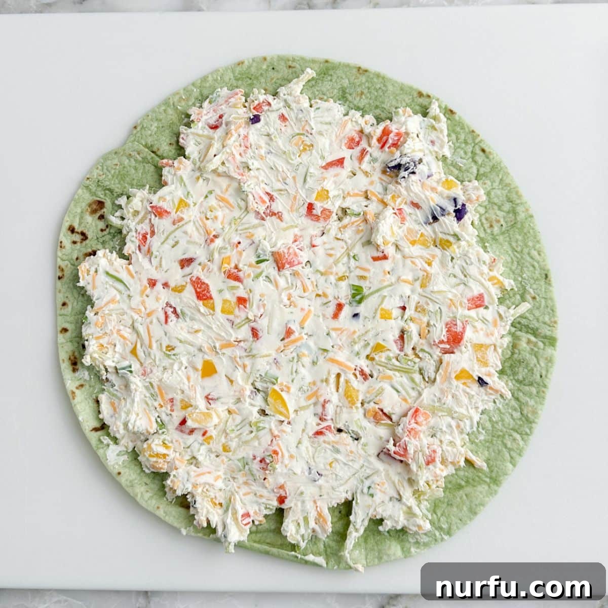 A green spinach tortilla with an even layer of cream cheese and vegetable mixture spread over its surface.