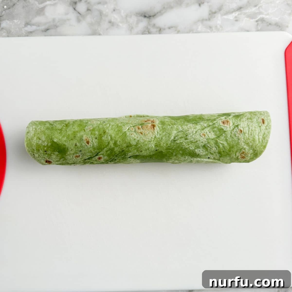 A tightly rolled green spinach tortilla filled with the cream cheese and vegetable mixture, resting on a cutting board.