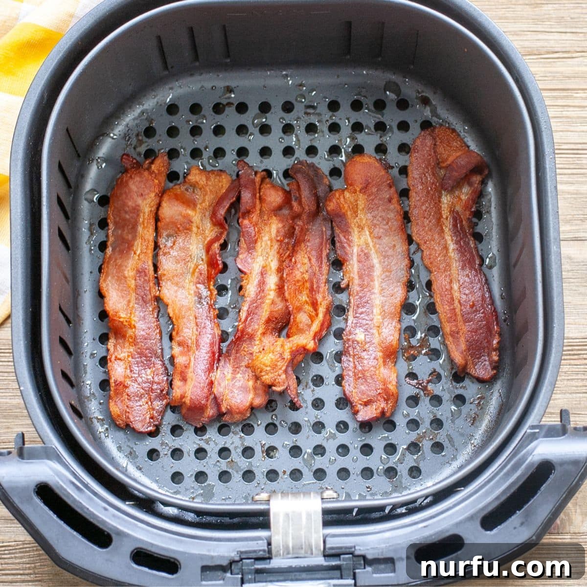 Crispy Air Fryer Bacon 3 Freshly cooked, golden-brown bacon slices resting on a paper towel-lined plate after being removed from an air fryer basket.