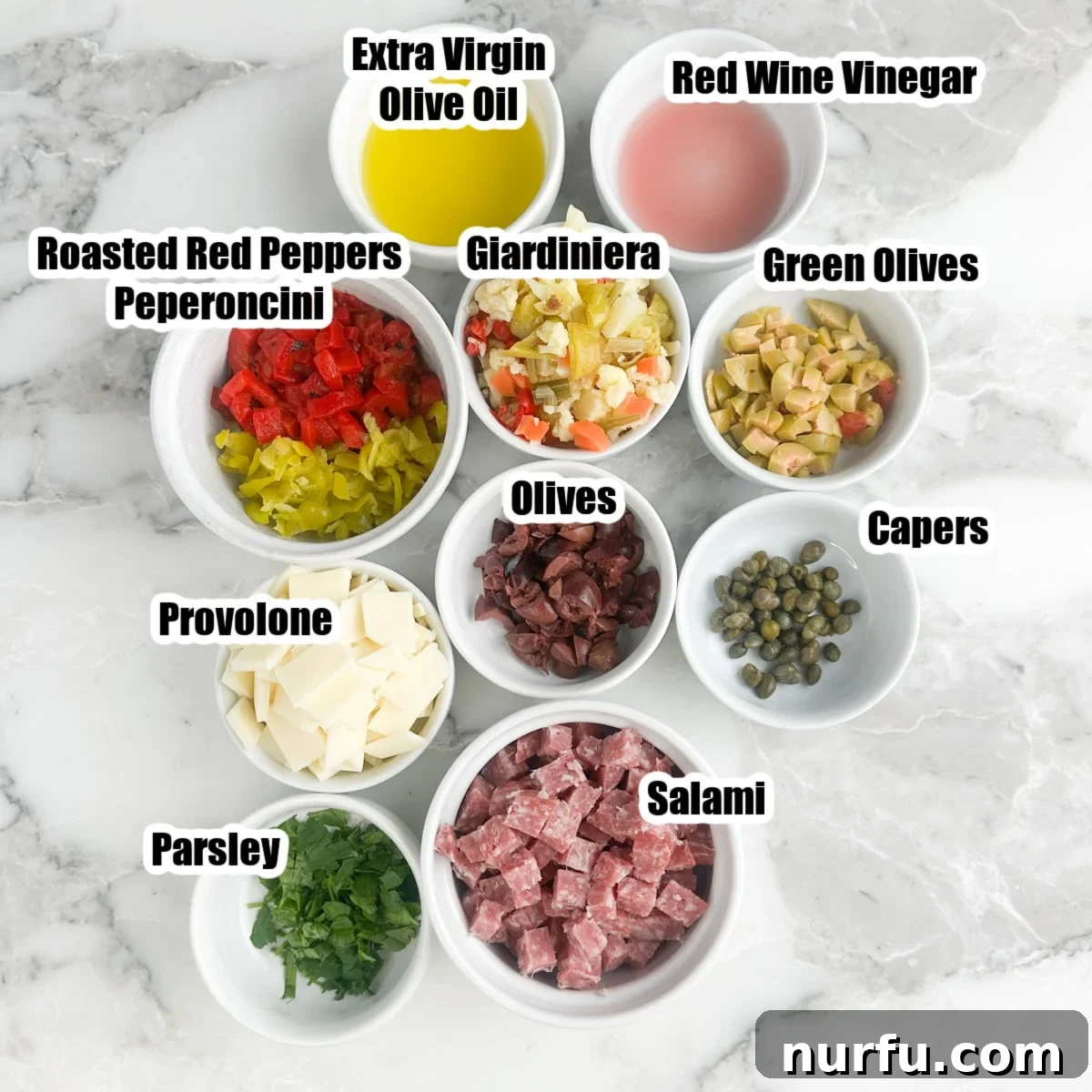 New Orleans Muffuletta Party Dip 3 A selection of fresh ingredients laid out for muffuletta dip, including diced salami, provolone cheese, green olives, kalamata olives, capers, giardiniera, peperoncini, roasted red peppers, fresh parsley, olive oil, and red wine vinegar.