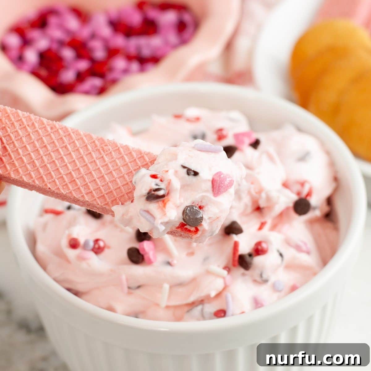 Pink Party Dip 2 A bowl of vibrant pink dessert dip, with a pink cookie partially submerged, ready for a delicious bite. The dip is adorned with colorful sprinkles, hinting at its festive appeal.