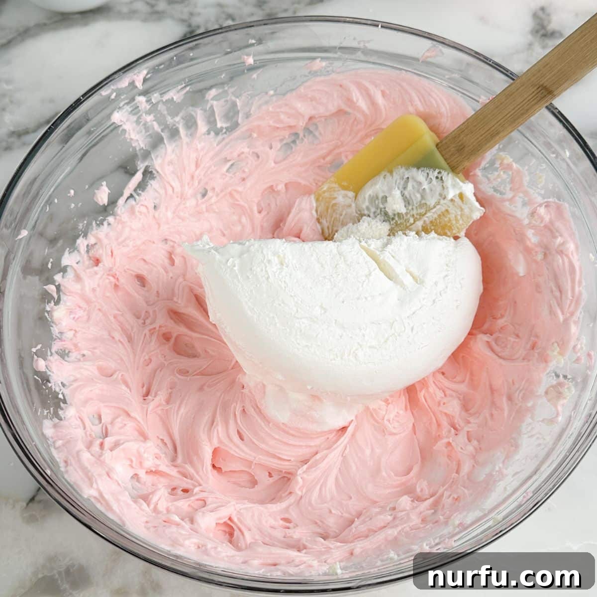 Pink Party Dip 5 A bowl of vibrant pink cream cheese mixture, with thawed Cool Whip being gently folded in using a rubber spatula, ensuring a light and airy consistency for the dessert dip.