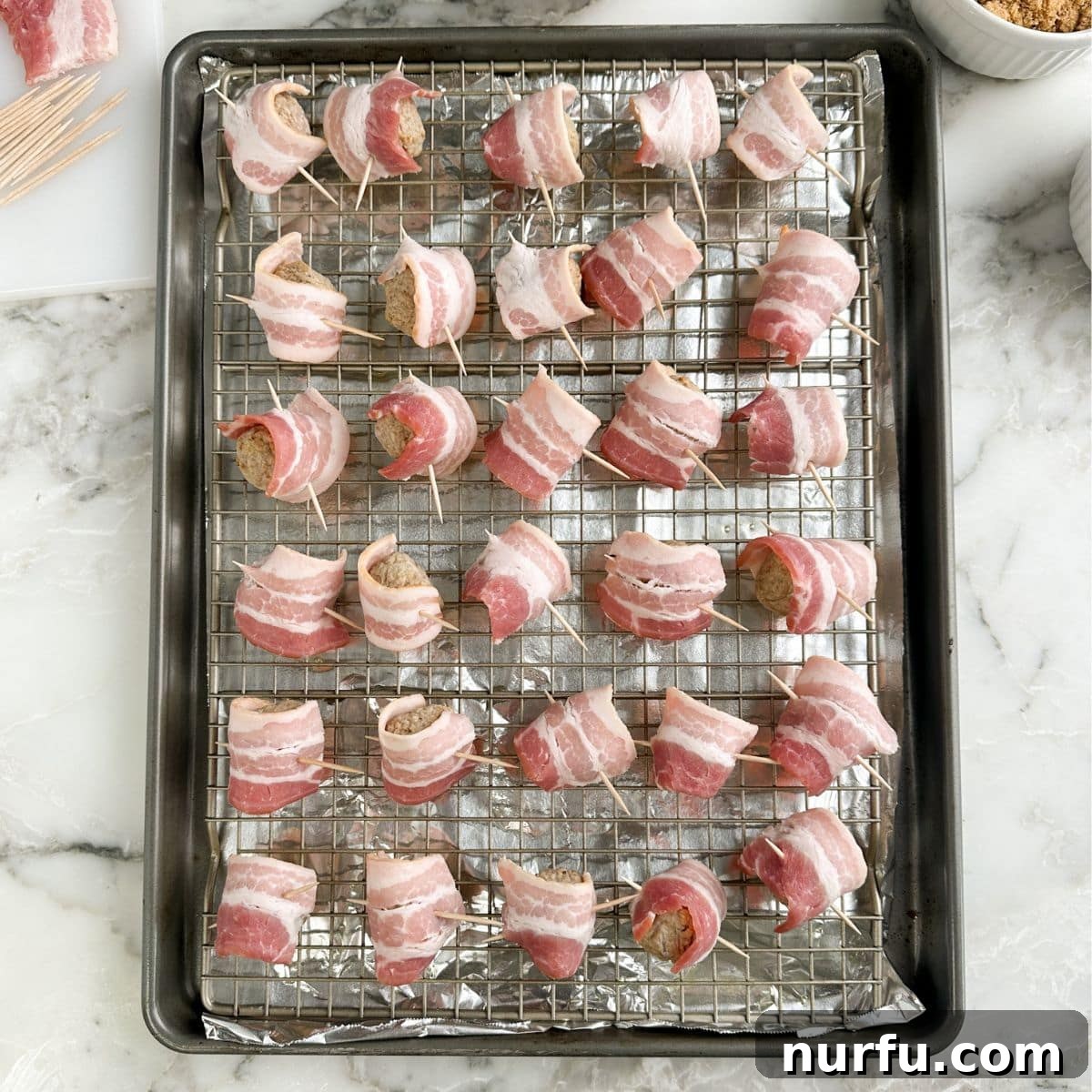 A baking sheet filled with uncooked bacon wrapped meatballs, each secured with a toothpick, before being placed in the oven.