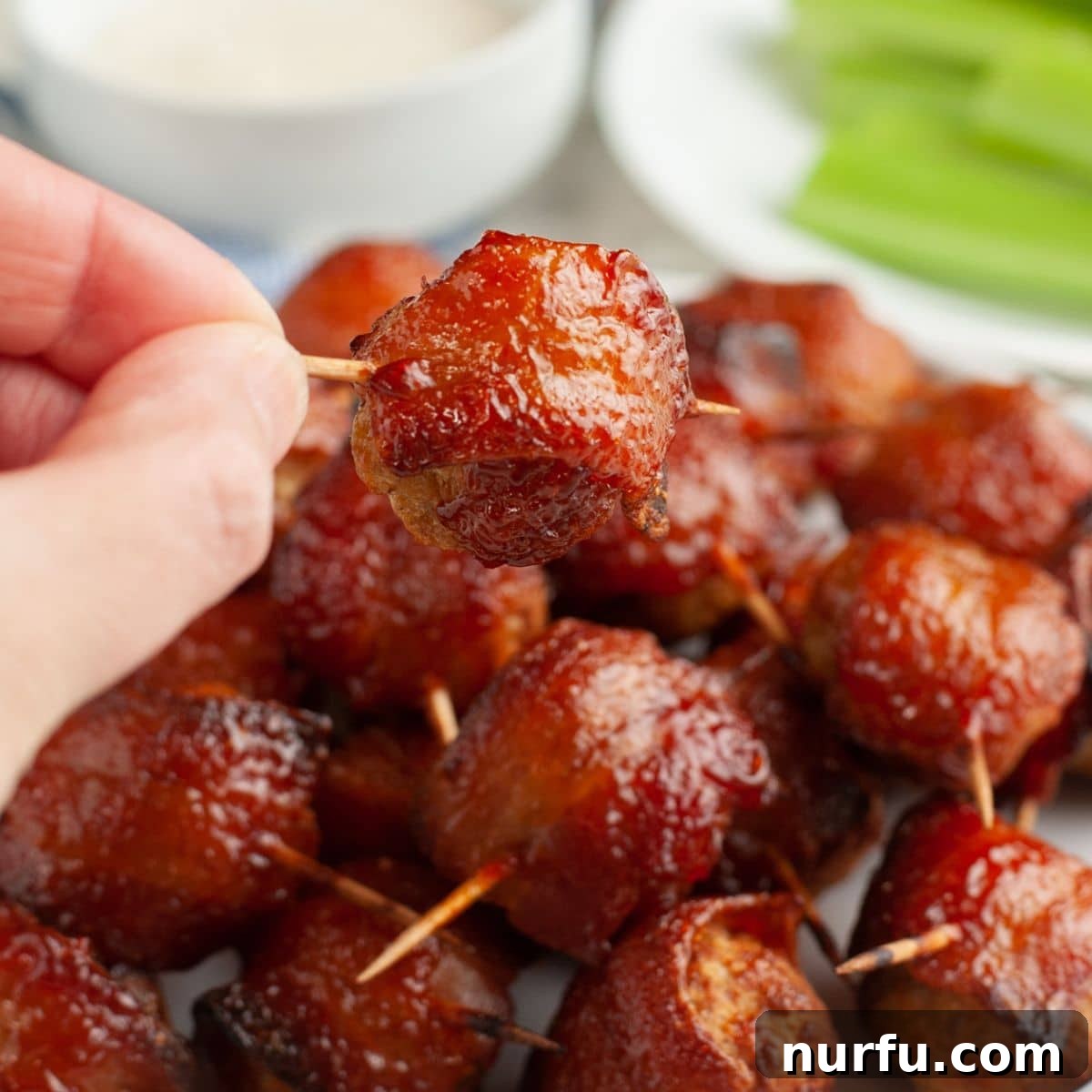 A hand holds up a single perfectly baked bacon wrapped meatball, showcasing its crispy bacon and glossy glaze, ready for a party.