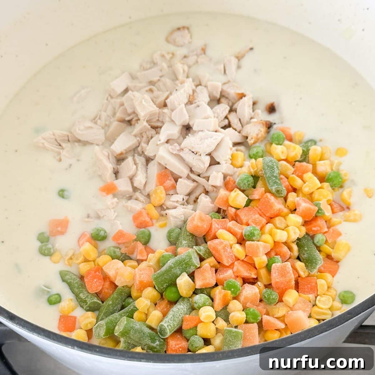 Hearty Chicken Pot Pie Pasta 7 A pot with a creamy sauce containing shredded chicken and frozen mixed vegetables, stirred together to combine the flavors before adding pasta.
