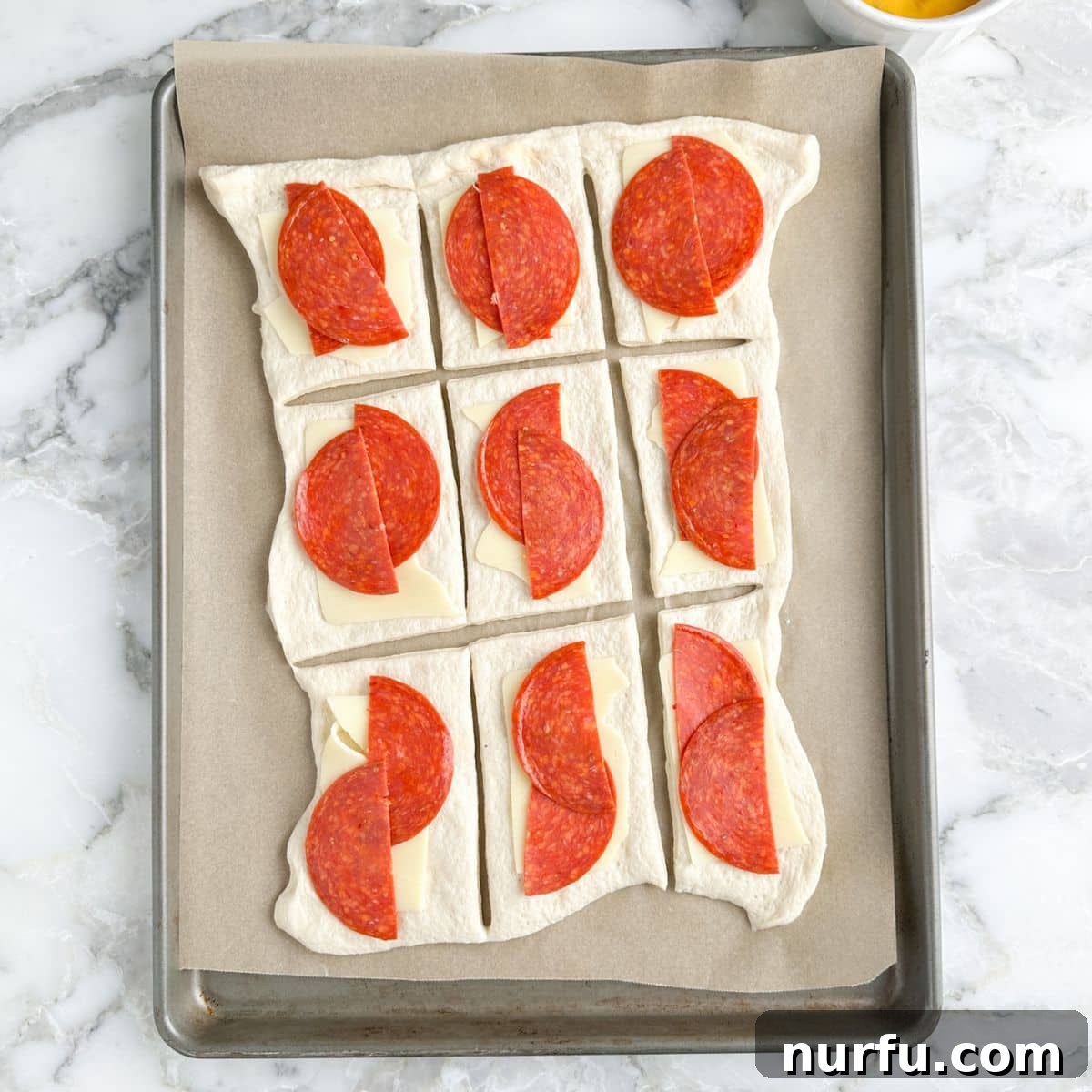 Cheesy Pepperoni Rolls 5 Unrolled bread dough sections on a baking pan, each topped with two halves of mozzarella cheese and two halves of pepperoni slices, ready for rolling.