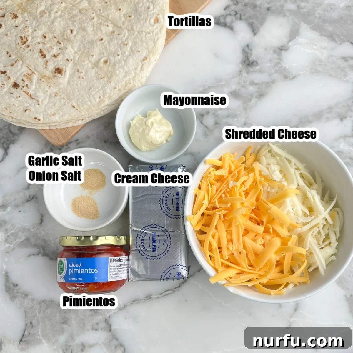 A collection of ingredients laid out: a bowl of shredded cheese, a jar of pimentos, a block of cream cheese, a jar of mayonnaise, a stack of tortillas, garlic powder, and onion powder.