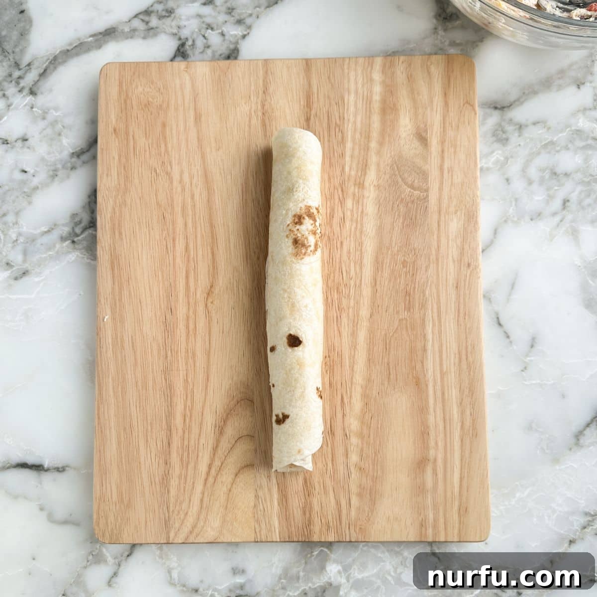 A tightly rolled tortilla filled with pimento cheese, wrapped in plastic wrap and placed on a wooden cutting board.