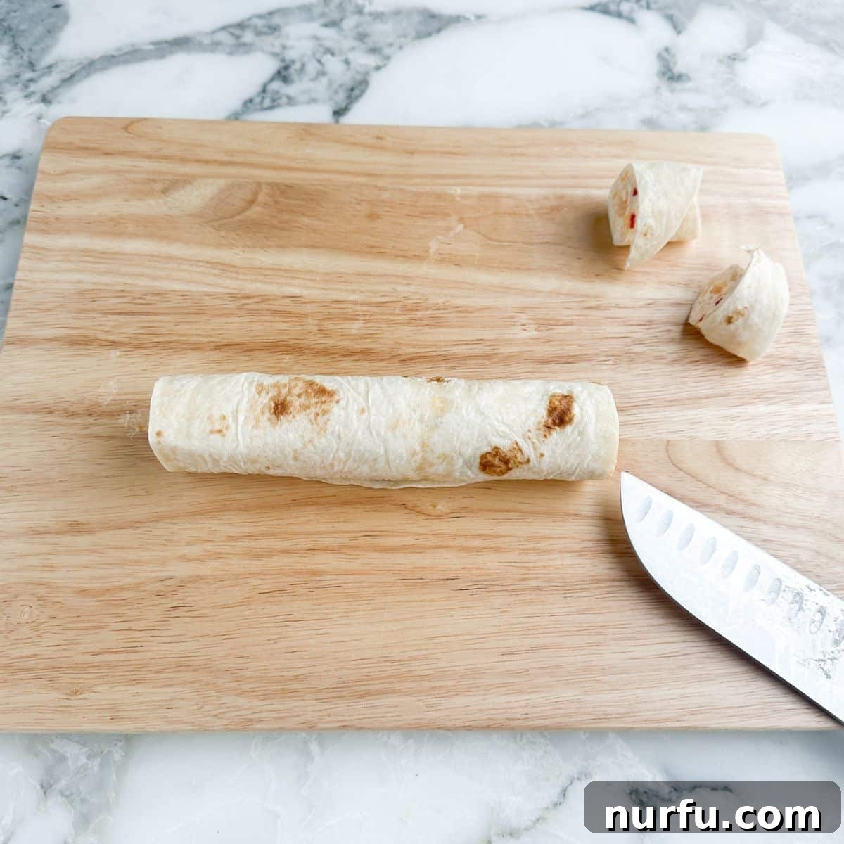 A cutting board with a rolled tortilla, a knife, and sliced pimento cheese pinwheels.