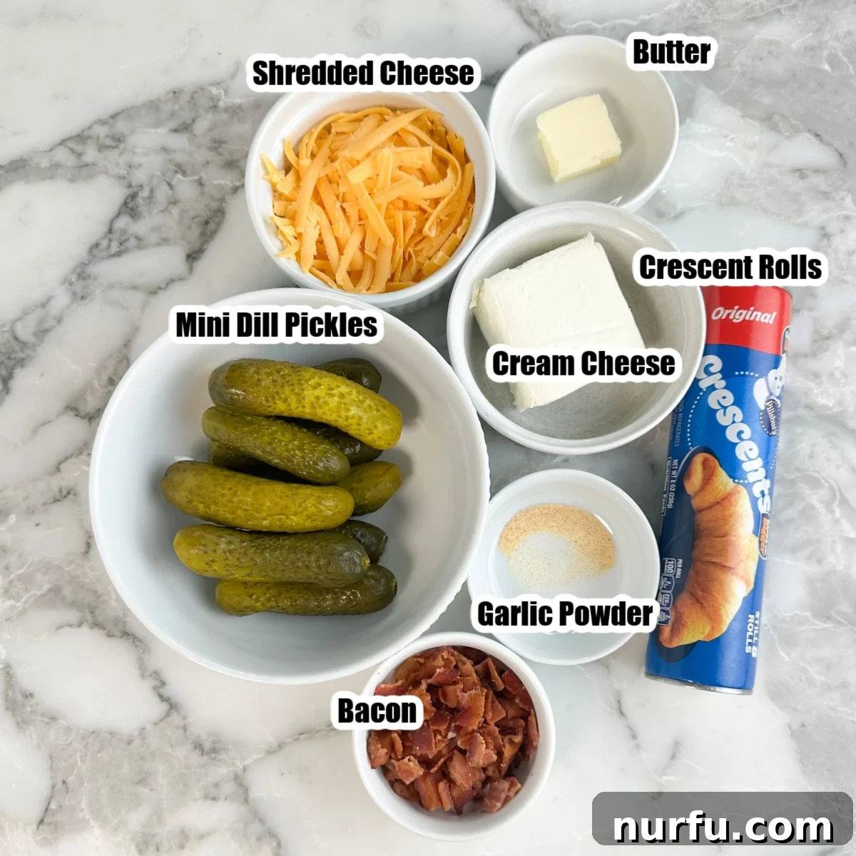 Tangy Pickle Bites 3 A collection of ingredients laid out: a bowl of baby dill pickles, shredded cheddar cheese, crumbled cooked bacon, softened cream cheese, garlic powder, melted butter, and a package of crescent roll dough.