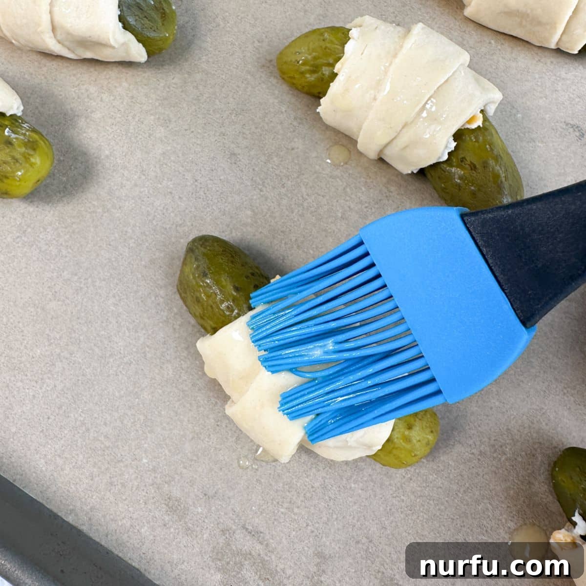 Tangy Pickle Bites 7 A hand brushing melted butter over the unbaked crescent roll pickles on a baking sheet.