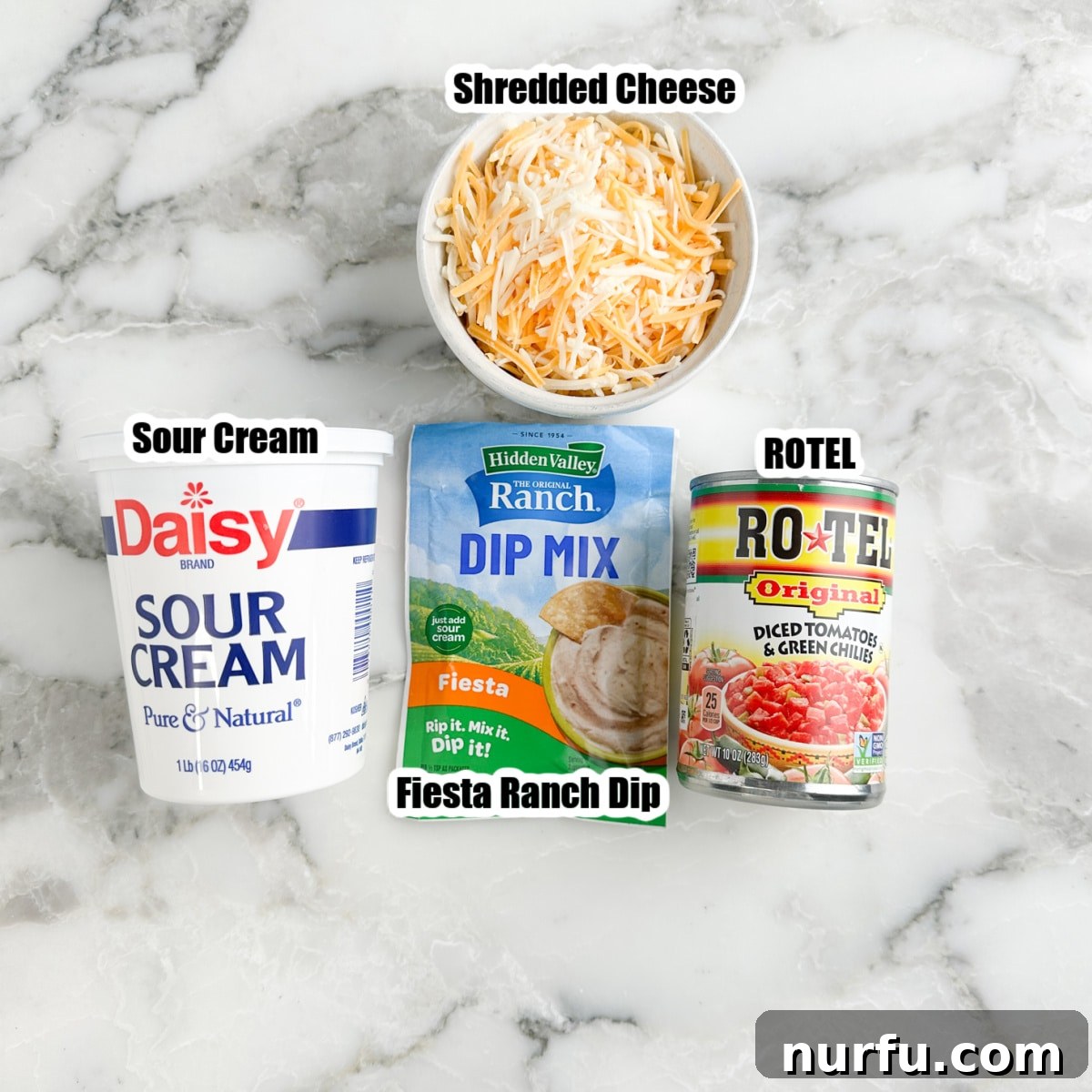 Zesty Fiesta Ranch Dip 3 A flat lay showing the four main ingredients for Fiesta Ranch Dip: a container of sour cream, a bowl of shredded Mexican cheese blend, a packet of Hidden Valley Fiesta Ranch Dip Mix, and a can of ROTEL diced tomatoes and green chilies.