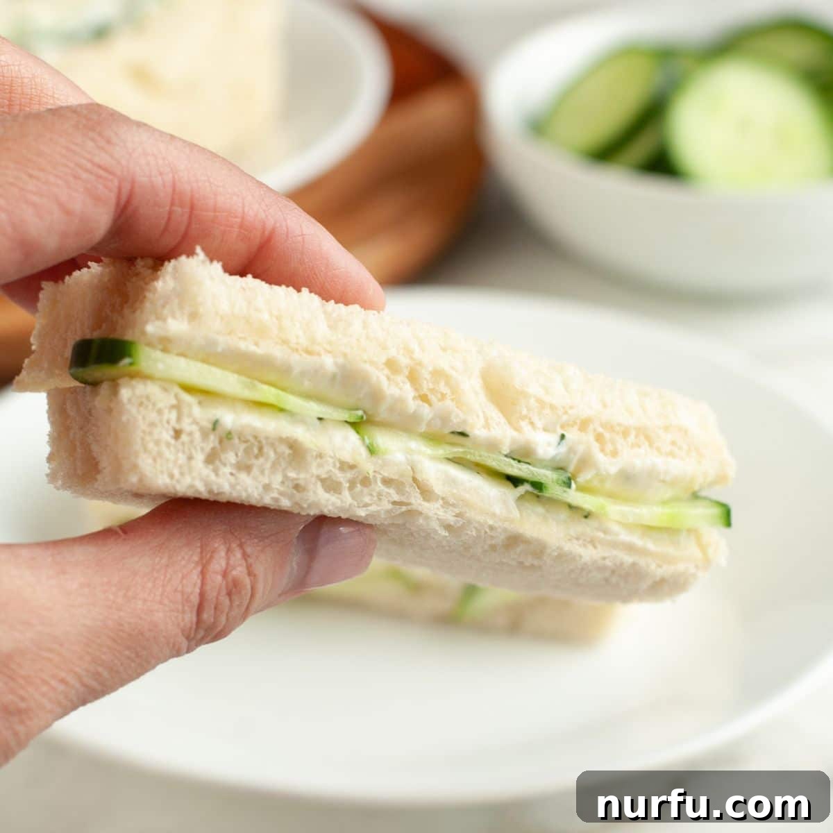 Elegant Cucumber Tea Bites 8 A hand holding a perfectly cut cucumber sandwich, showcasing its delicate layers.